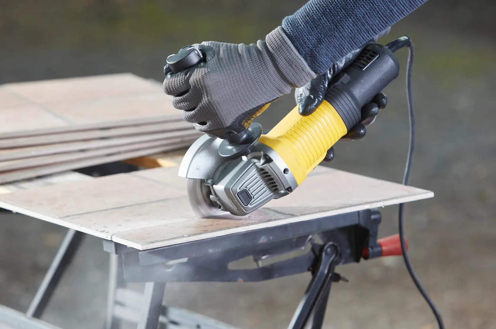 Photo of STANLEY FMEG210K angle grinder cutting tile, close-up with gloved hands and yellow handle.